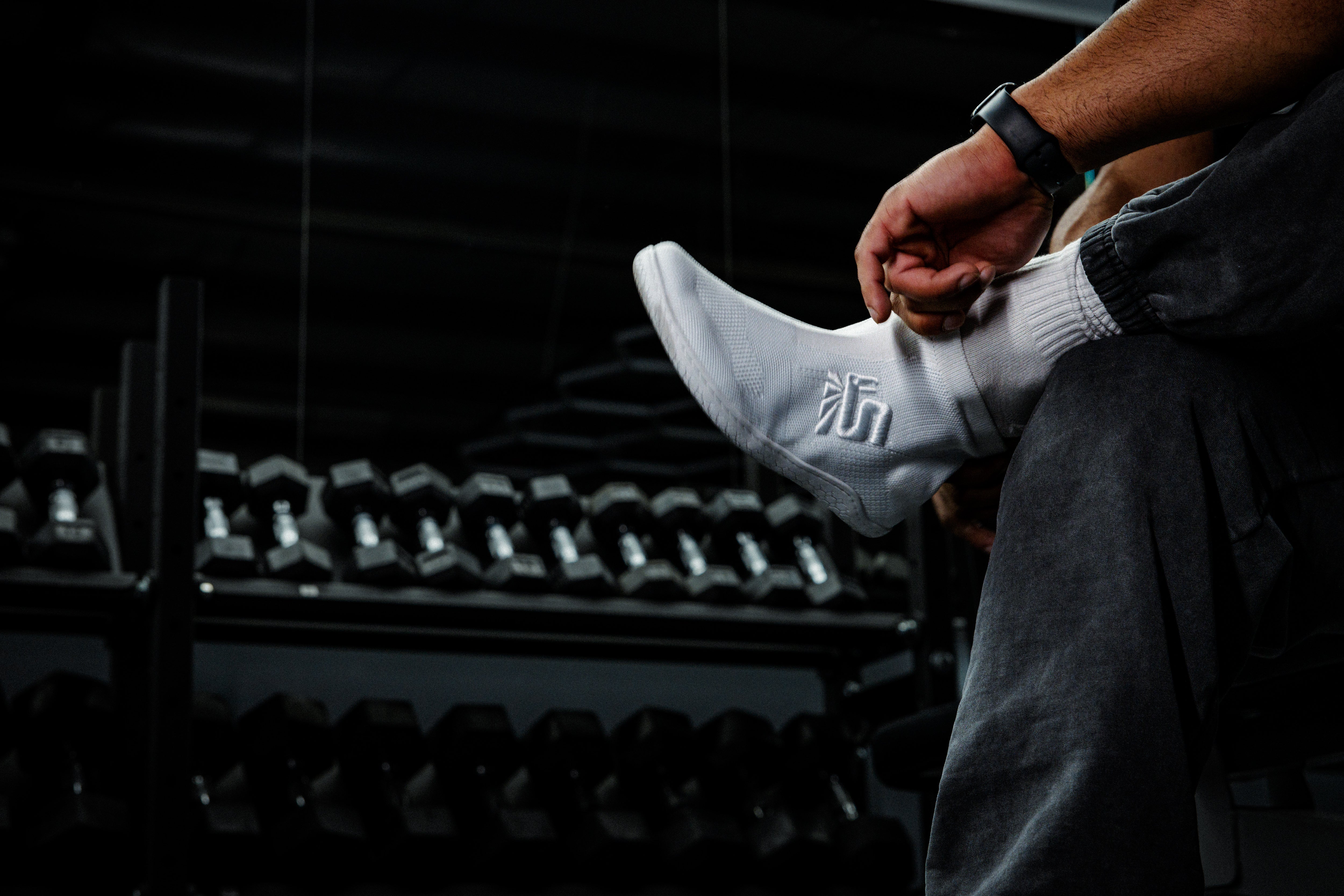 Putting on a white Otishi shoe with dumbbells in the background